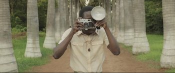 Movie still from “Palm Trees in the Snow” (2015), directed by Fernando González Molina – A man taking a picture with a camera in front of trees; Medium shot, Low angle