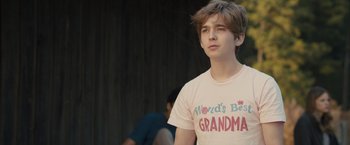 Movie still from “Paper Towns” (2015), directed by Jake Schreier – A young man wearing a t - shirt that says " world's best grandma "; Close Up shot, Over the shoulder angle