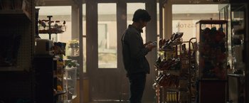Movie still from “Paper Towns” (2015), directed by Jake Schreier – A man standing in front of shelves of drinks looking at his cell phone; Medium shot, Over the shoulder angle