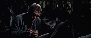 Movie still from “Papillon” (1973), directed by Franklin J. Schaffner – A man sitting on the ground with a cigarette in his mouth; Close Up shot, Low angle