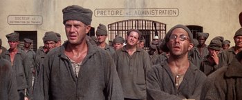 Movie still from “Papillon” (1973), directed by Franklin J. Schaffner – A group of men standing next to each other in front of a building; Medium shot, High angle
