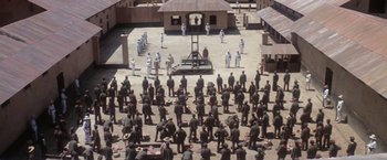 Movie still from “Papillon” (1973), directed by Franklin J. Schaffner – A group of people standing around a prison cell; Extreme Wide shot, High angle