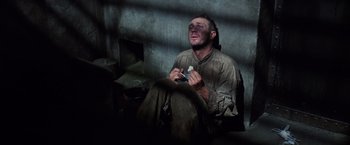 Movie still from “Papillon” (1973), directed by Franklin J. Schaffner – A man sitting in a dark room holding a knife; Medium shot, High angle