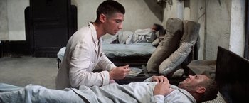 Movie still from “Papillon” (1973), directed by Franklin J. Schaffner – A man sitting on a bed while another man lies in bed; Medium shot, High angle