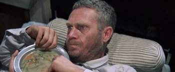Movie still from “Papillon” (1973), directed by Franklin J. Schaffner – A person sitting on a couch holding a bowl; Close Up shot, High angle