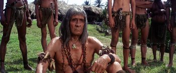 Movie still from “Papillon” (1973), directed by Franklin J. Schaffner – An old man with long hair and a beard is sitting on the grass with other men; Medium shot, Low angle