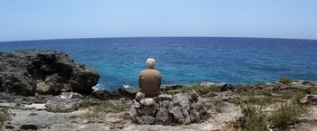 Movie still from “Papillon” (1973), directed by Franklin J. Schaffner – A man sitting on a rock looking out at the ocean; Wide shot, High angle