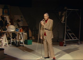 Movie still from “Parade” (1974), directed by Jacques Tati – A man in a suit and tie holding a microphone on a stage; Wide shot, Low angle