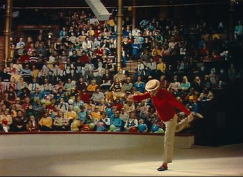 Movie still from “Parade” (1974), directed by Jacques Tati – A man in a red shirt and white hat is in front of an audience; Extreme Wide shot, High angle