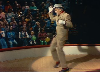 Movie still from “Parade” (1974), directed by Jacques Tati – A man in a suit and hat is on a stage in front of an audience; Wide shot, High angle