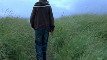 Movie still from “Paranoid Park” (2007), directed by Gus Van Sant – A man walking through tall green grass on a cloudy day; Medium shot, Low angle