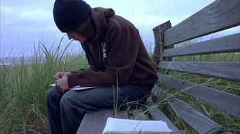Movie still from “Paranoid Park” (2007), directed by Gus Van Sant – A man sitting on top of a wooden bench writing; Medium shot, Low angle