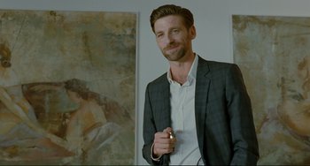 Movie still from “Passion” (2012), directed by Brian De Palma – A man in a suit holding a cigarette in his hand; Medium shot, Over the shoulder angle