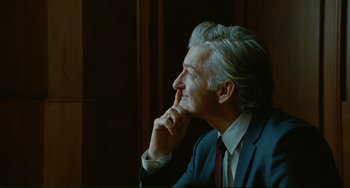 Movie still from “Passion” (2012), directed by Brian De Palma – An older man in a suit and tie sitting in front of a window; Close Up shot, Over the shoulder angle