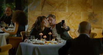 Movie still from “Passion” (2012), directed by Brian De Palma – A man and a woman sitting at a dinner table taking a picture with their cell phone; Medium shot, Over the shoulder angle
