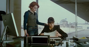 Movie still from “Passion” (2012), directed by Brian De Palma – A woman standing next to another woman at a desk; Medium shot, Over the shoulder angle