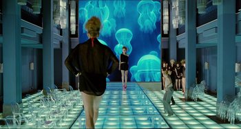 Movie still from “Passion” (2012), directed by Brian De Palma – A group of people standing on a runway in front of an aquarium; Wide shot, Over the shoulder angle