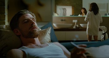 Movie still from “Passion” (2012), directed by Brian De Palma – A man laying in bed with a cigarette in his mouth; Medium shot, Over the shoulder angle