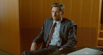 Movie still from “Passion” (2012), directed by Brian De Palma – A man in a suit and tie sitting in a chair; Medium shot, Over the shoulder angle