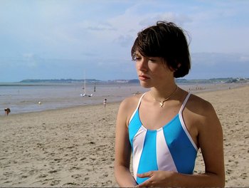 Movie still from “Pauline at the Beach” (1983), directed by Éric Rohmer – A woman in a blue and white bathing suit standing on a beach; Medium shot, High angle
