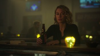 Movie still from “Peacemaker” (2022), created by James Gunn – A woman sitting at a table with a bottle of beer in front of her; Close Up shot, Over the shoulder angle
