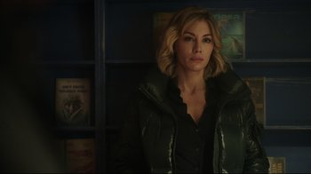 Movie still from “Peacemaker” (2022), created by James Gunn – A woman in a black jacket standing in front of a book case; Close Up shot, Over the shoulder angle