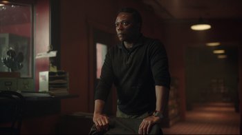 Movie still from “Peacemaker” (2022), created by James Gunn – A man in a black shirt is sitting down; Medium shot, Low angle