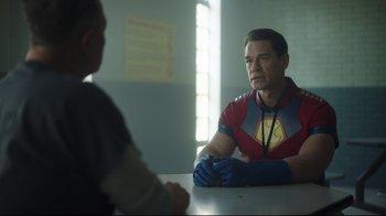 Movie still from “Peacemaker” (2022), created by James Gunn – A man in a red and blue shirt sitting at a table with another man; Medium shot, Over the shoulder angle