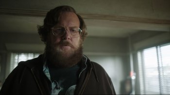 Movie still from “Peacemaker” (2022), created by James Gunn – A man with a goatee wearing glasses and a brown jacket; Close Up shot, Over the shoulder angle