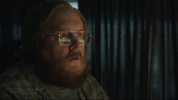 Movie still from “Peacemaker” (2022), created by James Gunn – A man with a goatee wearing glasses looking at the camera; Close Up shot, Low angle