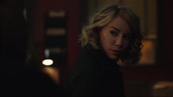 Movie still from “Peacemaker” (2022), created by James Gunn – A woman with blonde hair is standing in a dark room; Close Up shot, Over the shoulder angle