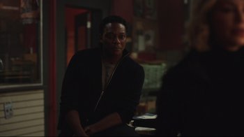 Movie still from “Peacemaker” (2022), created by James Gunn – A man sitting at a table in a dark room; Medium shot, Over the shoulder angle