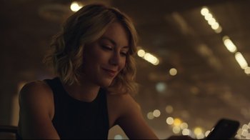 Movie still from “Peacemaker” (2022), created by James Gunn – A beautiful young lady sitting at a table in front of lights; Close Up shot, Over the shoulder angle