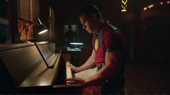 Movie still from “Peacemaker” (2022), created by James Gunn – A man in a red shirt is playing the piano; Medium shot, Low angle