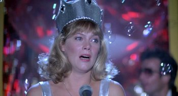 Movie still from “Peggy Sue Got Married” (1986), directed by Francis Ford Coppola – A woman wearing a crown and a microphone in front of a crowd; Close Up shot, Low angle