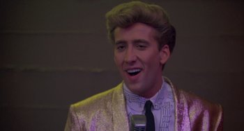 Movie still from “Peggy Sue Got Married” (1986), directed by Francis Ford Coppola – A man in a suit and tie holding a microphone in his hand; Close Up shot, Low angle