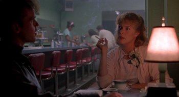 Movie still from “Peggy Sue Got Married” (1986), directed by Francis Ford Coppola – A woman sitting at a table smoking a cigarette; Medium shot, Over the shoulder angle