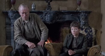 Movie still from “Pelle the Conqueror” (1987), directed by Bille August – A man and a boy sitting next to a fireplace; Medium shot, Over the shoulder angle