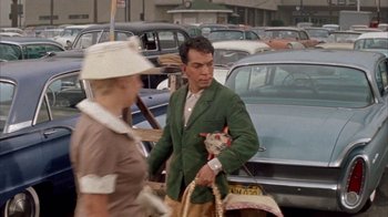 Movie still from “Pepe” (1960), directed by George Sidney – A man in a green jacket is walking in a parking lot while a woman in a white hat looks at him; Medium shot, High angle