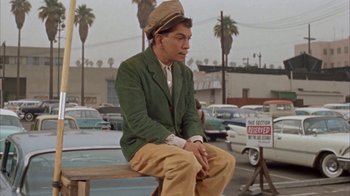Movie still from “Pepe” (1960), directed by George Sidney – A man sitting on top of a wooden bench in a parking lot; Medium shot, Low angle