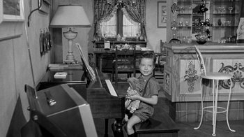 Movie still from “Period of Adjustment” (1962), directed by George Roy Hill – A young boy sitting at a piano holding a teddy bear; Wide shot, High angle