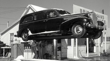 Movie still from “Period of Adjustment” (1962), directed by George Roy Hill – An old car is being lifted on a lift; Wide shot, Low angle