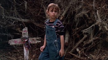Movie still from “Pet Sematary” (1989), directed by Mary Lambert – A little girl in overalls standing next to a pile of wood; Medium shot, High angle