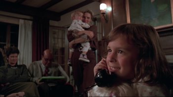 Movie still from “Pet Sematary” (1989), directed by Mary Lambert – A young girl talking on a phone while a man holds a baby in his arms; Medium shot, Low angle