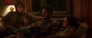 Movie still from “Pete's Dragon” (2016), directed by David Lowery – A man sitting on a couch looking at a book; Medium shot, Over the shoulder angle