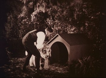 Movie still from “Peter Pan” (1924), directed by Herbert Brenon – A man and a dog in front of a doghouse in a wooded area; Wide shot, High angle
