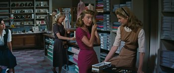 Movie still from “Peyton Place” (1957), directed by Mark Robson – A woman in a dress is looking at the camera while another woman in a dress is looking at a box; Medium shot, Over the shoulder angle