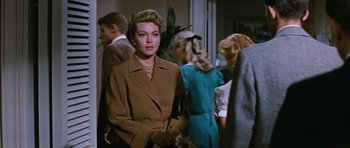 Movie still from “Peyton Place” (1957), directed by Mark Robson – A woman in a brown coat standing in front of a group of people; Medium shot, Over the shoulder angle