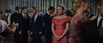 Movie still from “Peyton Place” (1957), directed by Mark Robson – A woman in a pink dress standing in front of a group of people; Medium shot, Over the shoulder angle