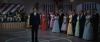 Movie still from “Peyton Place” (1957), directed by Mark Robson – A man standing in front of a group of people in formal wear; Wide shot, Low angle